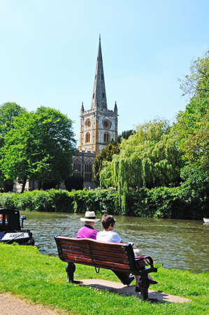 Stratford-upon-Avon, UK - May 18, 2014 - Holy Trinity Church seen across the River Avon with a couple sitting on a bench in the foreground, Stratford-Upon-Avon, Warwickshire, England, United Kingdom, Western Europe のeditorial素材
