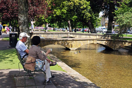 Bourton-on-the-Water, UK - June 12, 2014 - A couple sitting on a bench drinking tea along the River Windrush in the village centre, Bourton on the Water, Gloucestershire, England, UK, Western Europe のeditorial素材