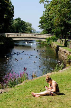 Tamworth, United Kingdom - July 23, 2014 - Young woman sitting on the riverbank alongside the River Anker using a mobile phone, Tamworth, Staffordshire, England, UK, Western Europe.のeditorial素材