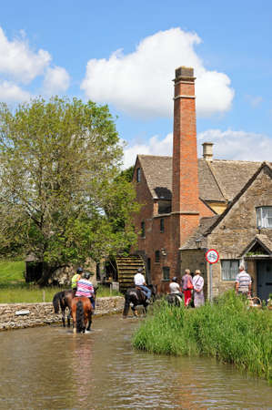 Lower Slaughter, UK - June 12, 2014 - Group of people riding horses along the River Eye with the old watermill to the rear, Lower Slaughter, Cotswolds, Gloucestershire, England, UK, Western Europe.のeditorial素材