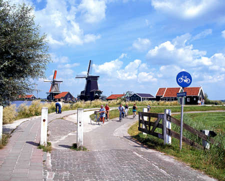 Row of windmills along the banks of the river Zaan with a cyclists in the foreground, Zaanse Schans, Holland, Netherlands, Europe.のeditorial素材
