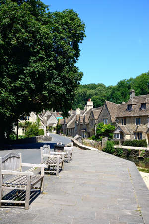 Stone cottages alongside the river Bybrook with wooden benches in the foreground, Castle Combe, Wiltshire, England, UK, Western Europe.のeditorial素材