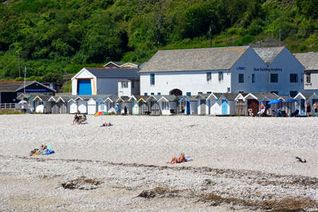 Holidaymakers on the shingle beach with beach huts and the Boat Building Academy to the rear, Lyme Regis, Dorset, England, UK, Western Europe.のeditorial素材