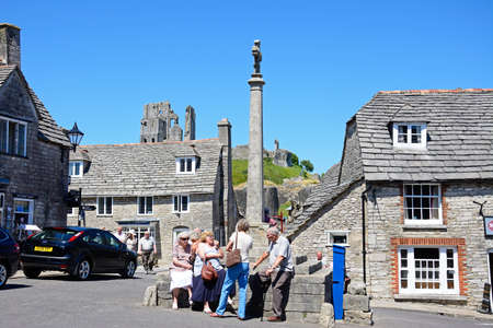 Tourists around the stone cross in the village centre with the castle to the rear, Corfe, Dorset,...