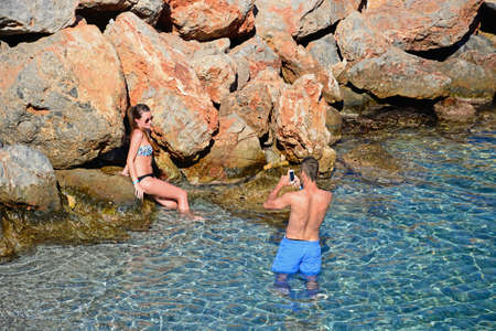 Man taking a photo of a woman against the rocks on his mobile phone, Agios Nikolaos, Crete, Greece, Europe.のeditorial素材