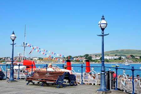 Tourists sitting on a wooden bench along the Victorian pier with views towards the town, Swanage, Dorset, England, UK, Western Europe.のeditorial素材