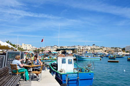 Traditional Maltese Dghajsa fishing boats in the harbour with waterfront buildings to the rear and two women relaxing at a restaurant on the quayside, Marsaxlokk, Malta, Europe.のeditorial素材