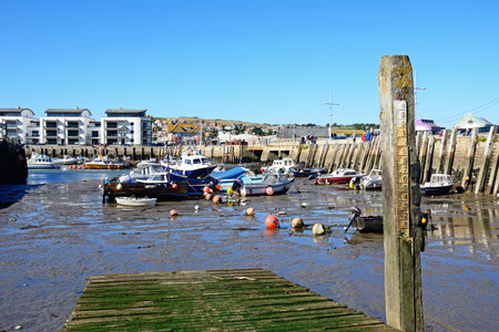 WEST BAY, UK - OCTOBER 10, 2022 - View of traditional fishing boats moored in the harbour at low tide with the town and countryside to the rear and a water depth gauge to the right, West Bay, Dorset, UK, Europe, October, 2022.のeditorial素材