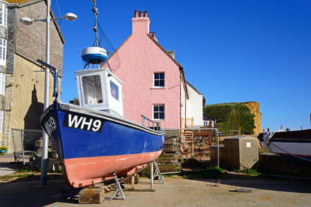 WEST BAY, UK - OCTOBER 10, 2022 - A traditional fishing boat in dry dock with pretty beachside cottages and Jurassic cliffs to the rear, West Bay, Dorset, UK, Europe, October, 2022.のeditorial素材
