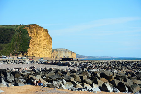 WEST BAY, UK - OCTOBER 10, 2022 - Couple with a dog sitting against the rocks on the beach with views towards the Jurassic Coast cliffs, West Bay, Dorset, UK, Europe, October, 2022.のeditorial素材