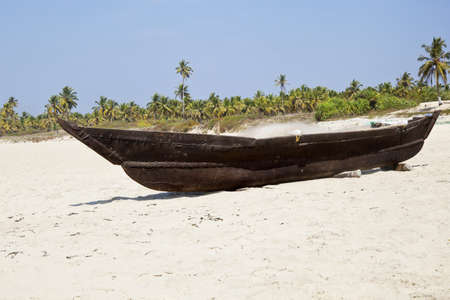 Horizontal landscape of a Goan fishermans wooden boat with his fishing nets and tackle on board resting on ramps on a white sandy beach with a coconut palm tree lined shoreline  の写真素材