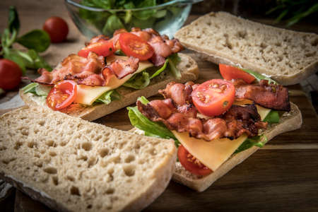 Ciabatta sandwich with arugula salad, bacon and yellow cheese on wooden board.の写真素材