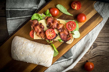 Ciabatta sandwich with arugula salad, bacon and yellow cheese on wooden board. Top view.の写真素材