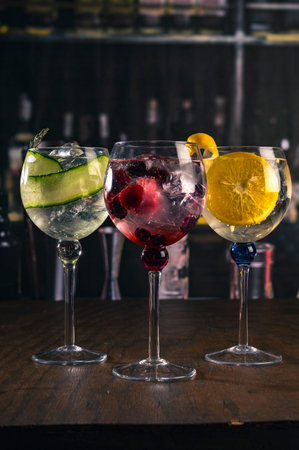 Three elegant gin and tonic cocktails with garnishes of orange, cucumber, and red berries on a rustic wooden bar, with a blurry bar background and moody lighting, perfect for mixologyの写真素材