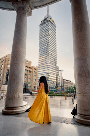 Elegant middle-aged woman in yellow dress exploring Mexico City near Torre Latinoamericana, captured by Palacio de Bellas Artes with modern skyline and historic sculpture in the background.の写真素材