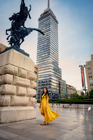 Elegant middle-aged woman in yellow dress exploring Mexico City near Torre Latinoamericana, captured by Palacio de Bellas Artes with modern skyline and historic sculpture in the background.の写真素材