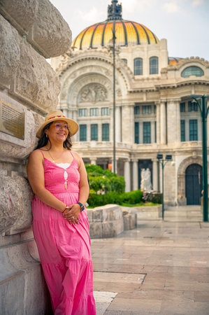 Happy woman wearing a pink dress and straw hat leaning against a stone wall near the Palacio de Bellas Artes in Mexico City, with the iconic golden dome and art deco architecture in background.の写真素材