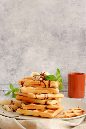 Hot, fresh stack of waffles with banana and chocolate sauce and a terracotta cup on grey background with copy spaceの写真素材