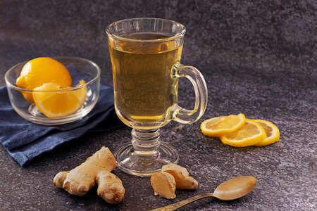 A glass of hot ginger and lemon tea with slices of lemon and ginger root on dark background