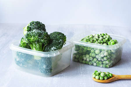 Frozen vegetables. Frozen broccoli and peas in plastic boxes and a wooden spoon on gray tableの写真素材