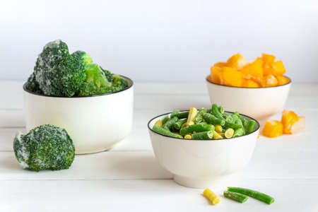 Frozen vegetables assortment. Frozen broccoli, French beans and pumpkin in white bowls on white tableの写真素材
