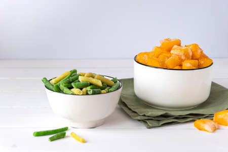 Frozen vegetables assortment. Frozen French beans and pieces of pumpkin in white bowls on white tableの写真素材