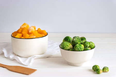 Frozen vegetables assortment. Frozen Brussels sprouts and pieces of pumpkin in white bowls on white tableの写真素材