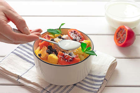 Bowl of fresh fruit salad with coconut flakes, cut tamarillo and yoghurt in the background. Ladys hand putting yoghurt into the salad.の写真素材