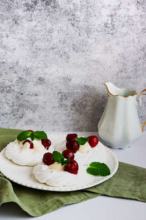 Meringue cakes decorated with whipped cream, cherries and mint leaves on a white plate, with a milk jug, side view with copy spaceの写真素材