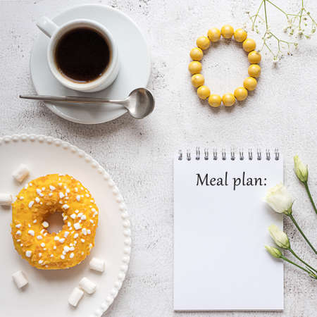 A blank notepad sheet with breakfast setting on gray concrete background. A cup of coffee, donut, flowers and a wooden bracelet. Concept of a lady's meal plan. Copy space.の写真素材