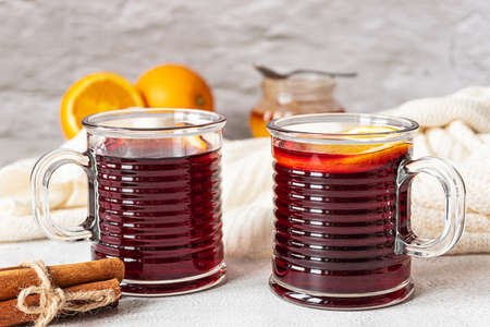 Two cups of hot mulled red wine with cinnamon sticks and orange. Spices, a jar of honey and a woolen knitted blanket. Gray concrete background.の写真素材