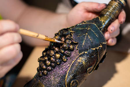 Female artist is using a painting brush on brown handmade bottle with leaves and grape made of coffee beansの写真素材