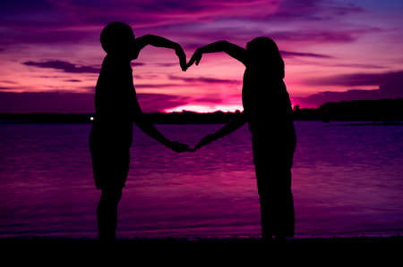 young couple making heart shape with arms on beach against pink light at sunsetの写真素材