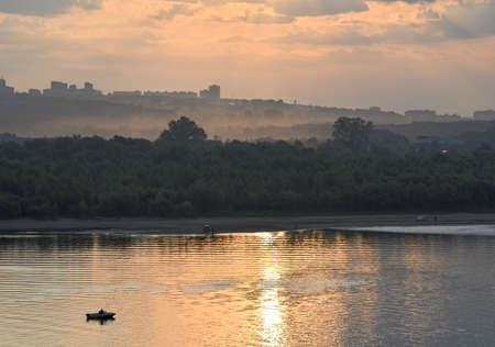 Landscape with a boat in the morning rays. Morning sky with Golden and gloomy clouds over the Bank of a wooded river in the morning fog. The water reflects the Golden light, the fisherman's boat. Novosibirsk region, Siberia, Russia, 2019の写真素材