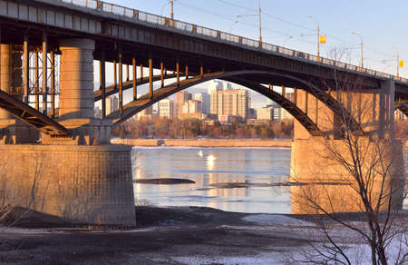 Arch of the Oktyabrsky automobile bridge over the Ob river. Powerful pillars, tall houses on the shore on the horizon in the morning light. Russia, Siberia, Novosibirskの写真素材