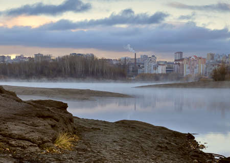 Novosibirsk foggy morning on the rocky Bank of the Ob river. Intertwining banks, houses and trees are reflected in the water. Autumn vegetation. The sky in gray cloud. Siberia, Russia, 2018の写真素材