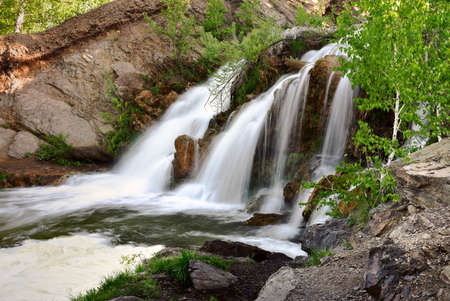 Belovsky waterfall in the spring. A small waterfall among the rocks, surrounded by birch trees with spring foliage in the rays of the sun. White streams of water, desolation, fresh greenery, stones, the nature of Siberia. Novosibirsk region, Russia, 2020の写真素材