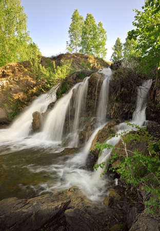 Belovsky waterfall in spring. A small waterfall among the rocks, surrounded by birch trees with spring foliage in the rays of the sun. White streams of water, without people, nature of Siberia. Novosibirsk region, Russia, 2020の写真素材