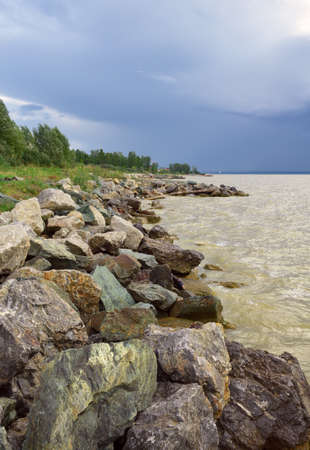 Big rocks on the Bank of the Novosibirsk reservoir, grass and trees, waves on the water, dark blue sky with clouds. Natural landscape. Novosibirsk region, Siberia, Russia, 2020の写真素材