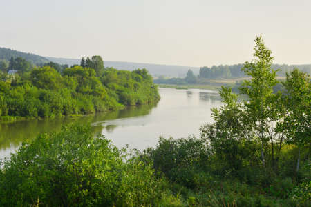 The winding riverbed, the surface of the water with reflections, green lush trees and shrubs on the...