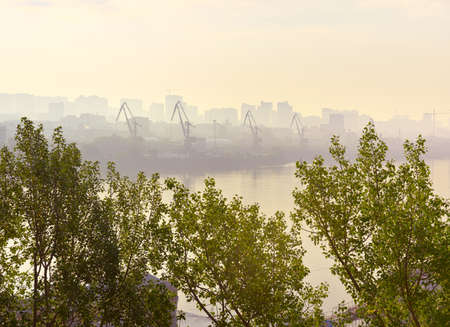 A foggy morning on the Ob River in Novosibirsk. The shore of a big city with port cranes in a yellow haze on the horizon behind the crowns of trees. Siberia, Russiaの写真素材