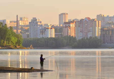 Novosibirsk, Siberia, Russia - 05/10/2019: Fisherman with a fishing rod knee-deep in the water at dawn. Morning on the Ob river, at home on the Bank of Novosibirsk in the morning sun, reflection in the water. Fishing is a favorite leisure of Russian menのeditorial素材