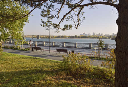 Novosibirsk, Siberia, Russia - 08.05.2019: Tree on the green lawn of the Park on the waterfront, bike path, lights, people walking, arched bridge and tall houses on the horizon in the eveningのeditorial素材