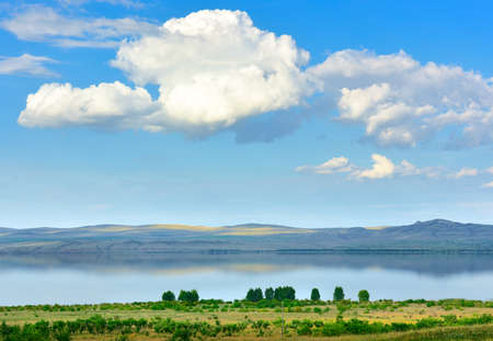 The shore of a steppe reservoir among blue mountains in summer under a big white cloud in a blue...