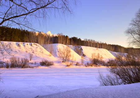 The bank of the Inya River in winter. A high slope with trees at the top among blue drifts of snow in the morning light against a clear sky. Novosibirsk, Siberia, Russiaの写真素材