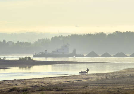 Novosibirsk region, Siberia, Russia - 08.20.2020: A fisherman walks along the Bank of the Ob river. A cargo barge sails through the morning fogのeditorial素材