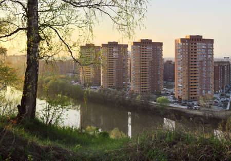 Birch with fresh spring foliage on the high Bank of a small river, high multi-storey red houses in the distance, Golden sky. Novosibirsk, Siberia, Russia, 2020の写真素材