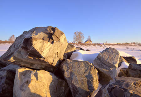 Stones on the shore of the Ob Sea. A snowy riverbank in spring, a yellow house and bare trees on a...