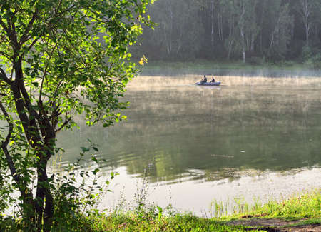 Novosibirsk region, Siberia, Russia - 09.20.2020: Two fishermen with a rod and oars on a rubber boat on the surface of a forest lake early in the morning. The mist over the water, the trees, the leaves on the branches, active rest on natureのeditorial素材