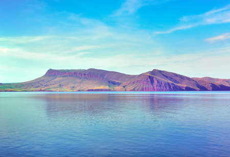 A green shore under a blue sky, red rocks in the distance. Krasnoyarsk Territory, Siberia, Russia